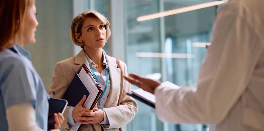 Medical professionals and a woman in a blazer discussing documents in a hospital corridor.