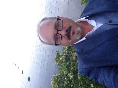 Man in glasses and suit outdoors near greenery and building.