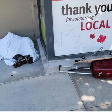 Person sleeping under a white sheet on the sidewalk near a 'Thank you for supporting local' sign.