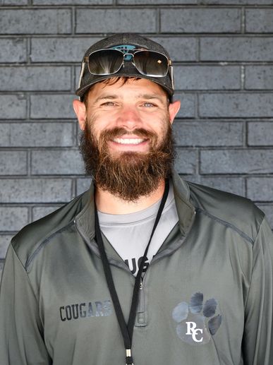 Bearded man in a cap and sunglasses smiling against a brick wall.