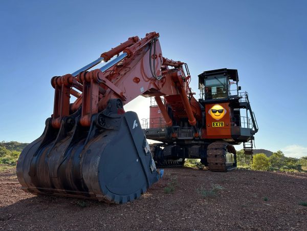 Large orange excavator with a cool emoji on the cabin, sitting on rocky ground.