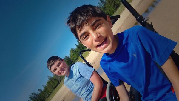 Two boys smiling outdoors on a sunny day near a sandy area and greenery.