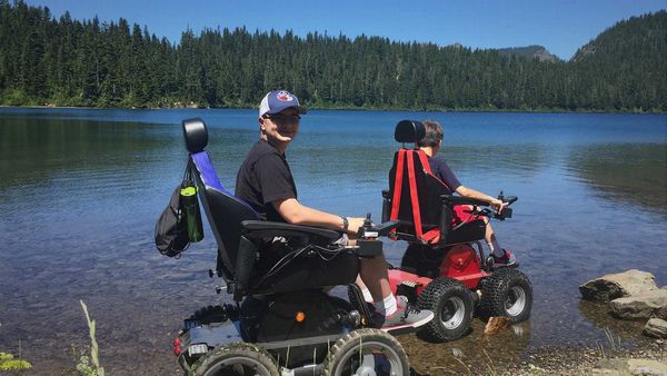 Two boys in motorized wheelchairs by a lakeside on a sunny day.