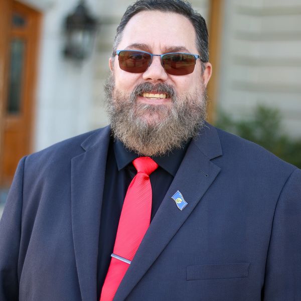 John J Looney Sr., in professional attire with a black suit, red tie, and sunglasses, stands confide