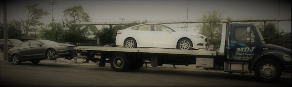 Tow truck carrying a white car and towing a gray car on the street.