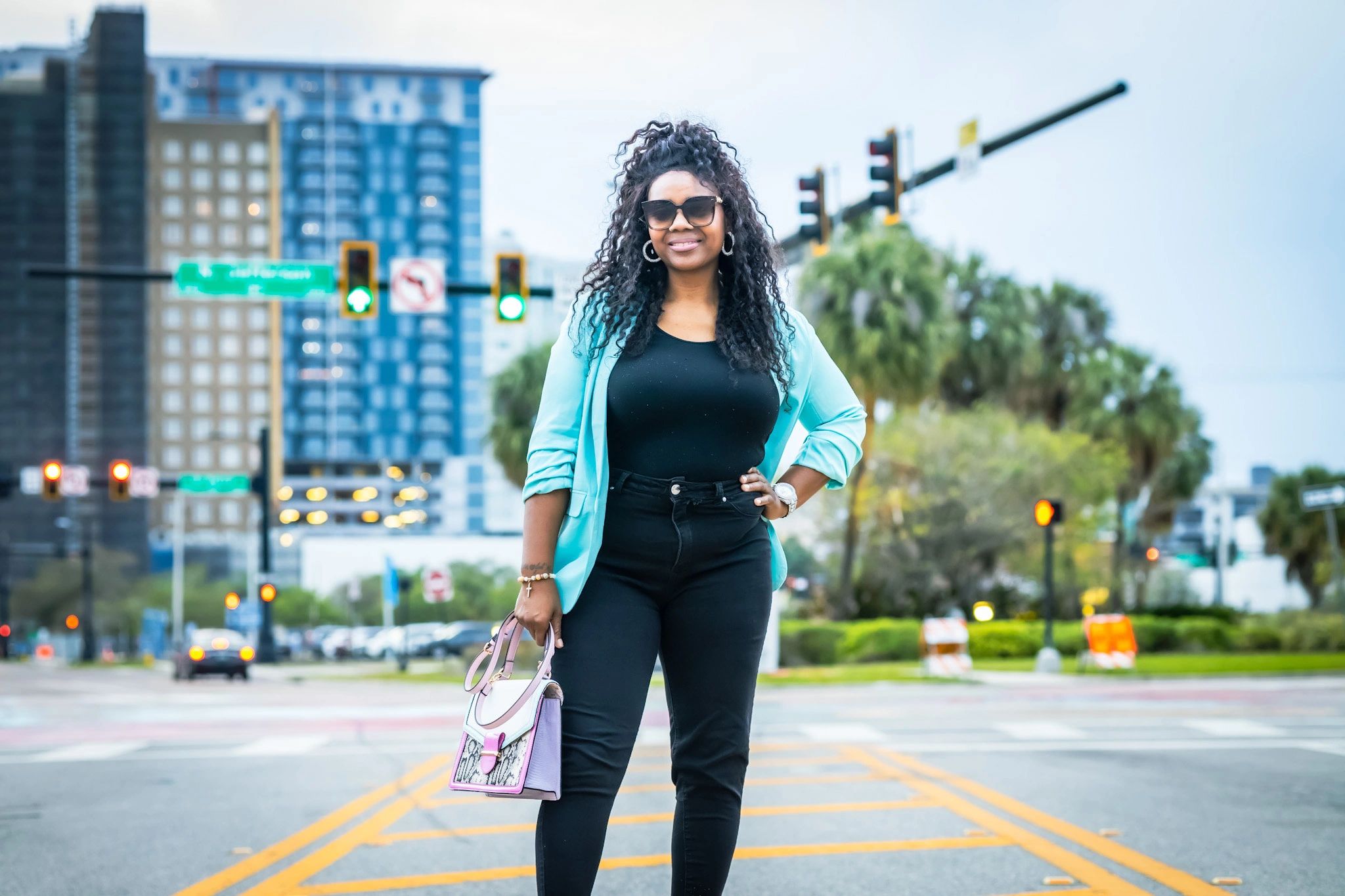 Confident woman standing in the city street wearing sunglasses and holding a handbag.
