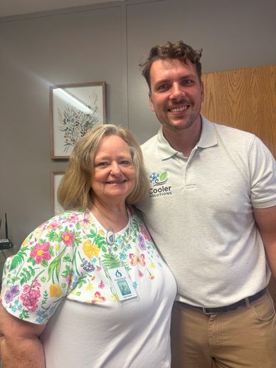 Two smiling people standing close indoors, one wearing a floral shirt and the other a white polo.
