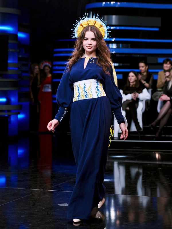 Model walks runway in a navy dress with embroidered corset and floral headpiece.