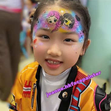Smiling child with colorful face paint featuring two cartoon characters on forehead.