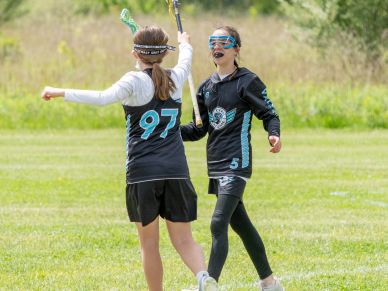 Two girls in lacrosse gear celebrating on a grassy field.
