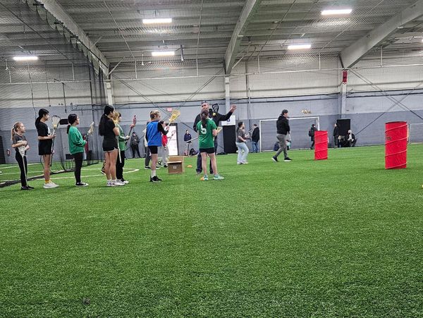 Indoor lacrosse practice with players lined up and coach instructing.