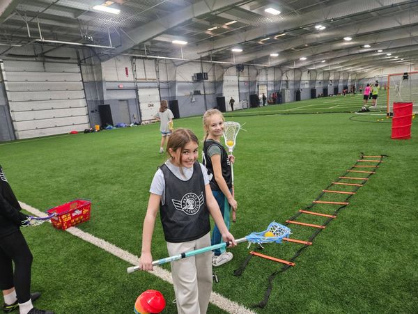 Two girls holding lacrosse sticks in an indoor sports facility.