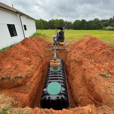 Excavator placing a large black tank into a deep trench near a white building.