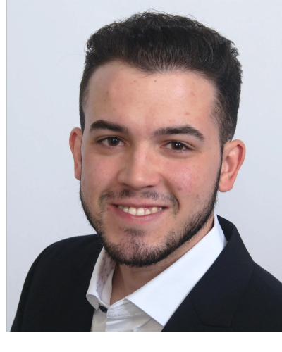 Young man with dark hair and beard in a black suit smiling.