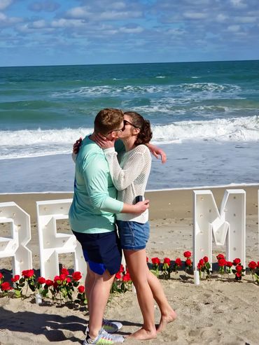 Couple kissing on the beach with roses and large white letters behind them.