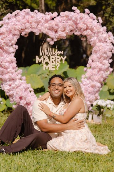 Couple embraces in front of a floral heart with 'Will You Marry Me?' sign.