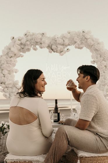 Couple smiling under a floral heart arch with a 'Will You Marry Me?' sign at the beach.