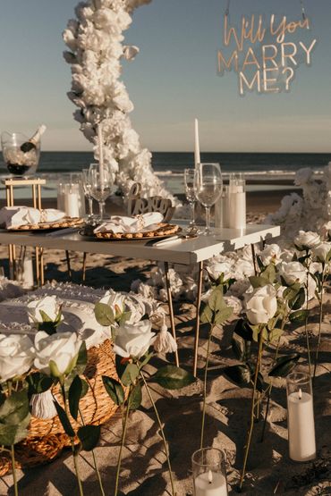 Romantic beach proposal setup with white roses and a "Will You Marry Me?" sign.