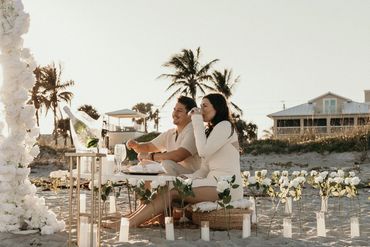 Couple enjoying a romantic beach picnic surrounded by white roses and candles.