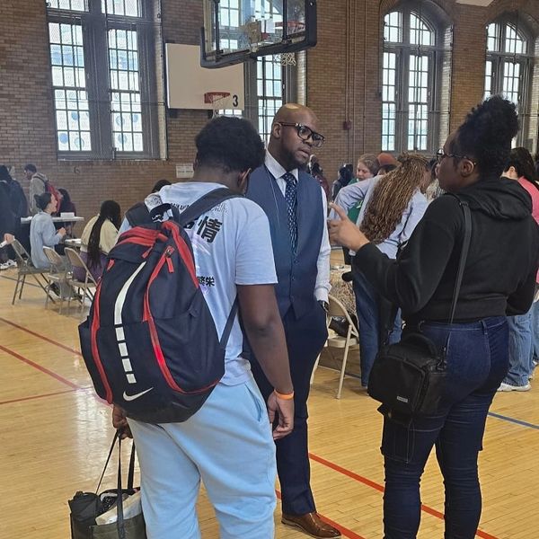 Three people engaged in conversation inside a gymnasium with basketball hoops and large windows.