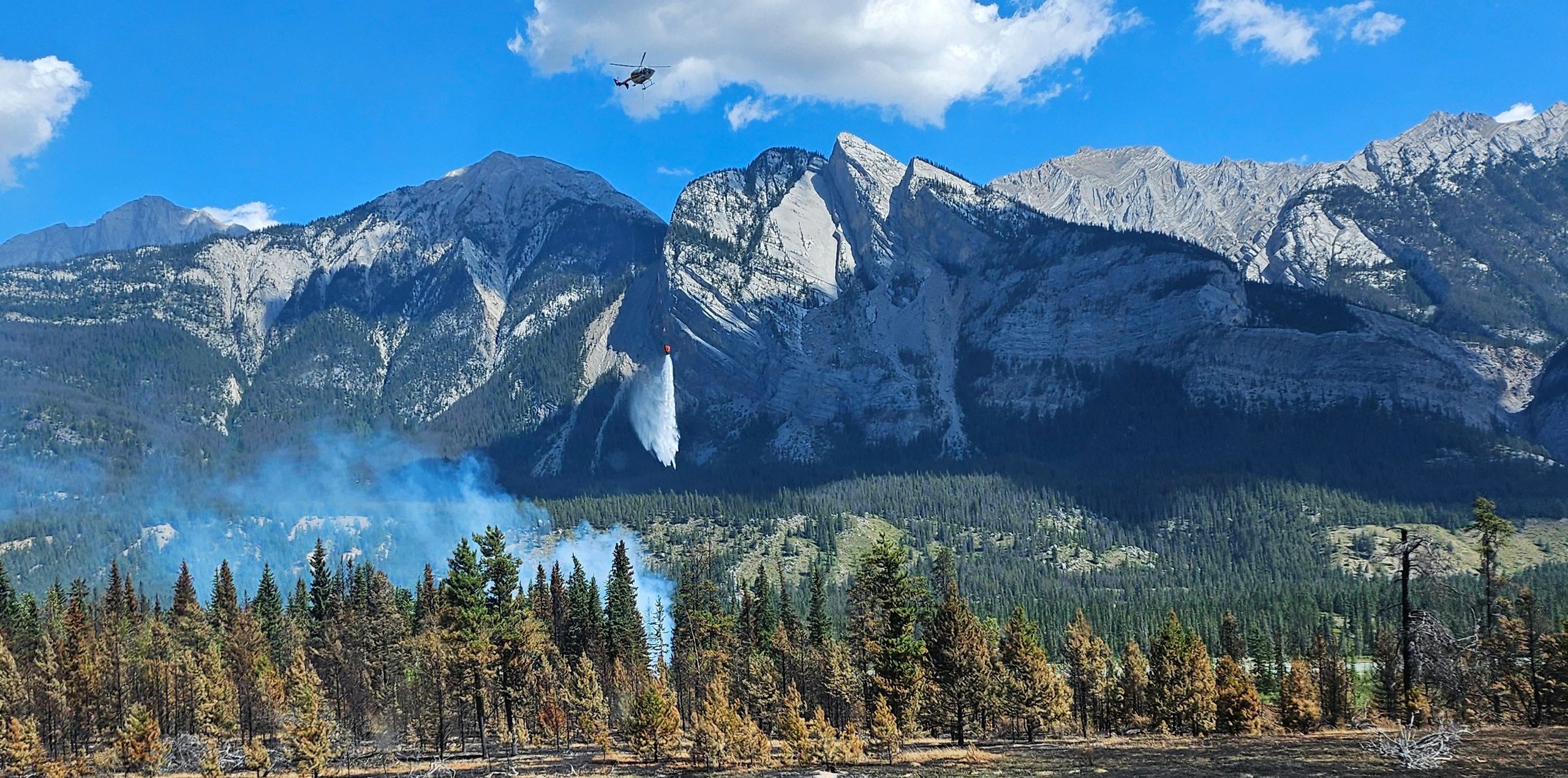A helicopter drops water to fight a forest fire in mountainous terrain.