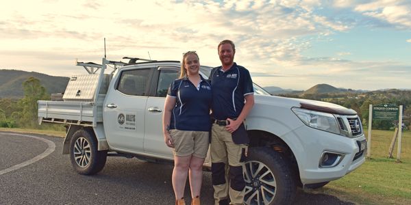 Couple standing in uniform in front of work ute, backdrop of Stroud Commons, mountains in distance.
