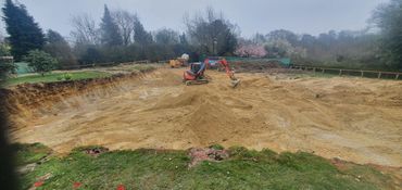 Excavator digging a large pit in a garden area surrounded by trees.