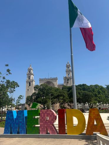 Colorful MÉRIDA sign in front of historic cathedral and Mexican flag under clear blue sky.