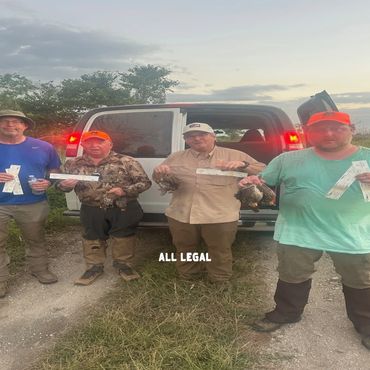 Four men holding hunting permits and game birds, standing in front of a van at dusk.