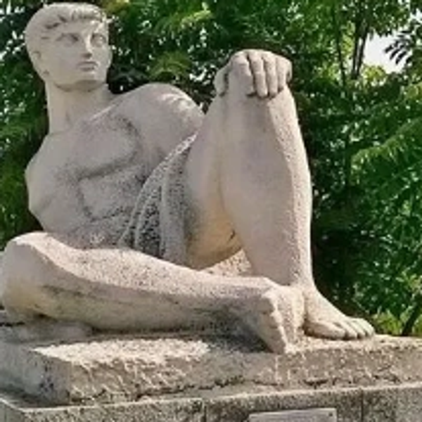 Stone sculpture of a seated man with a muscular build in an outdoor setting.