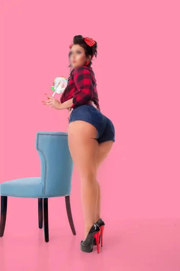 Woman in high heels and shorts posing with a lollipop and blue chair against pink background.