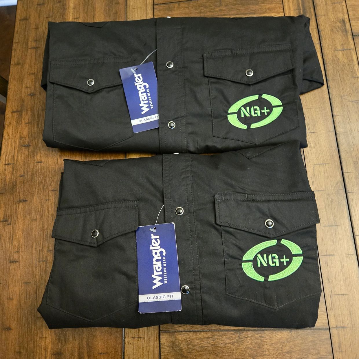 Two black Wrangler shirts with NG+ logo folded on a wooden table.
