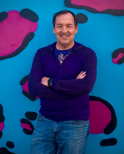 Smiling man with crossed arms stands before a colorful graffiti wall.