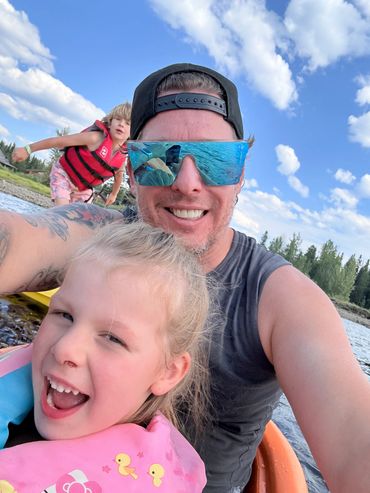 Child and adult smiling for a sunny lakeside selfie, both wearing sunglasses.