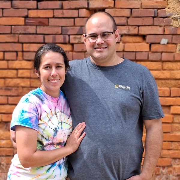 Ashlin and his wife, standing closely together in front of a textured, historic red brick wall.