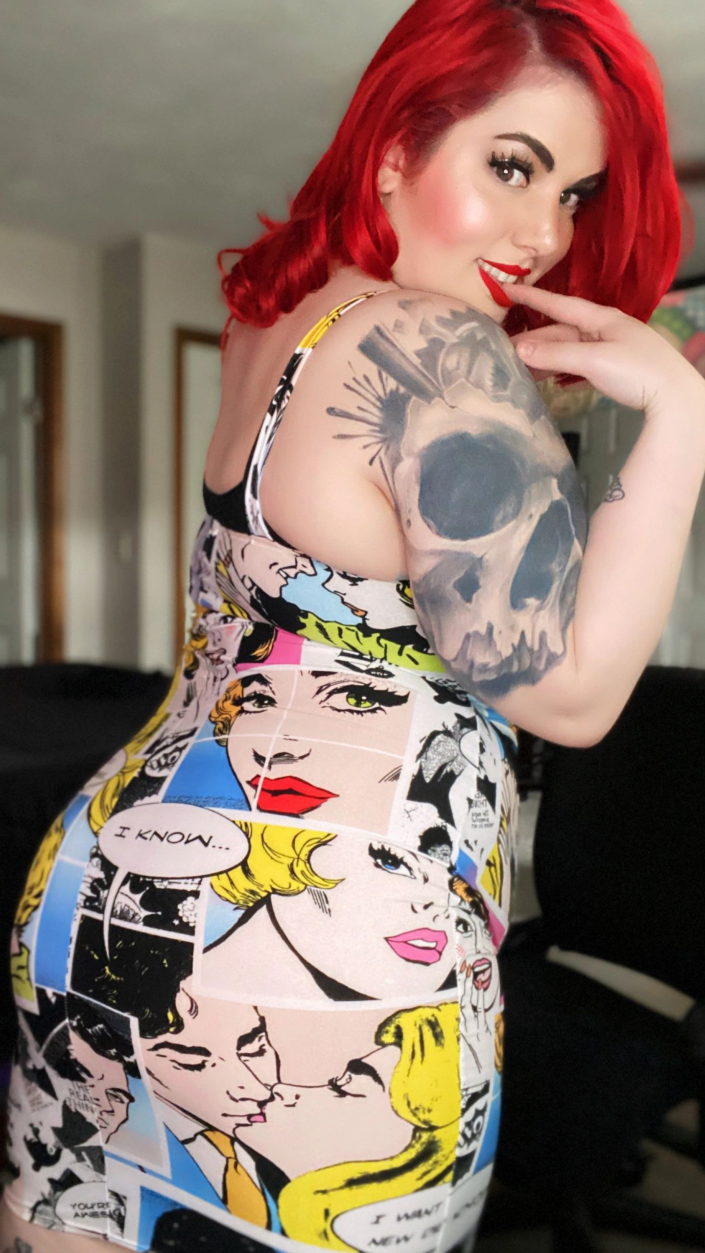tattooed redhead female pin up style model smiles and bites her finger wearing a comic book dress