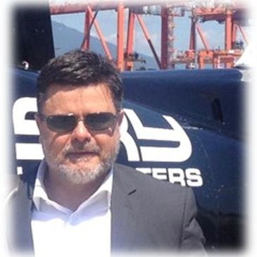 Man in sunglasses and blazer standing near a helicopter.
