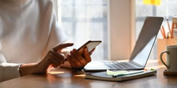 Hands using a cell phone while at a desk and in front of a laptop, notepad, and mug.