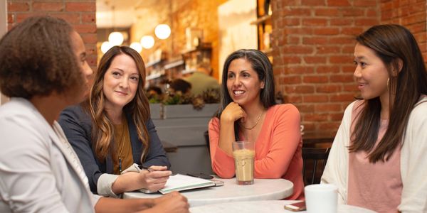 4 diverse women talking while sitting at two small round tables.
