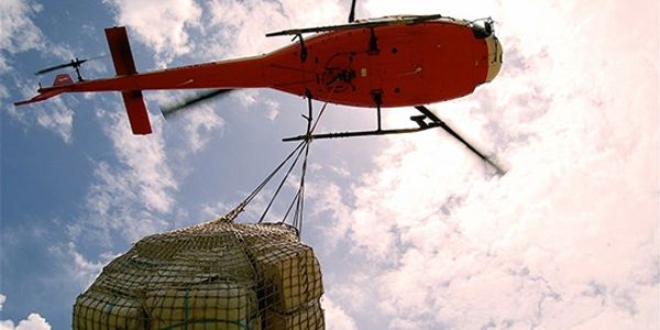 Red helicopter carrying cargo suspended in a net against a blue sky.