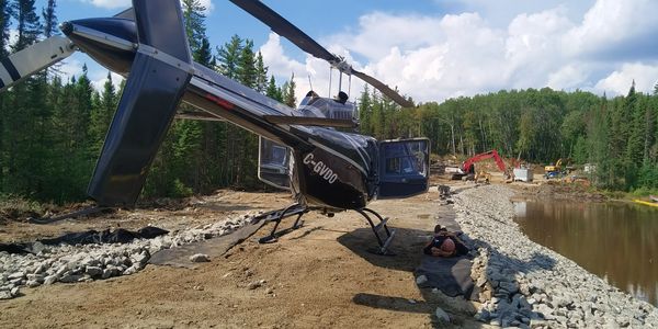 Helicopter landed on a dirt path near a river with construction equipment in the background.