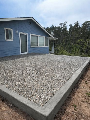 New pebble stone patio next to a blue house with forest backdrop.