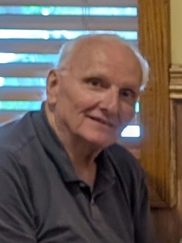 Elderly man smiling gently indoors with wooden blinds in the background.