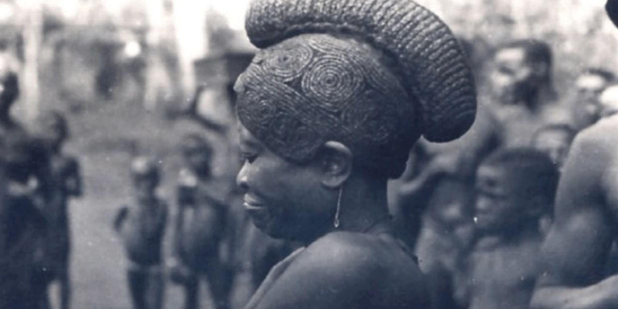 Profile of a woman with intricate braided hairstyle and traditional earring, surrounded by children in a blurred background.