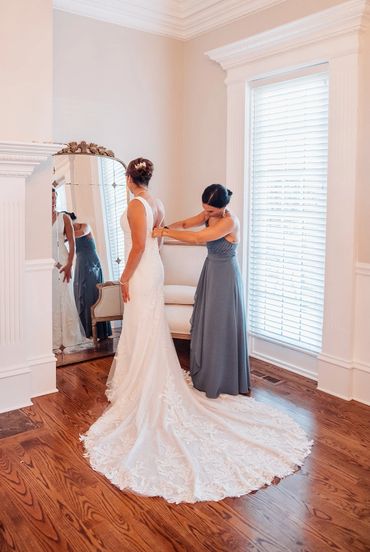 Bride getting her wedding dress adjusted by a bridesmaid in a bright room.