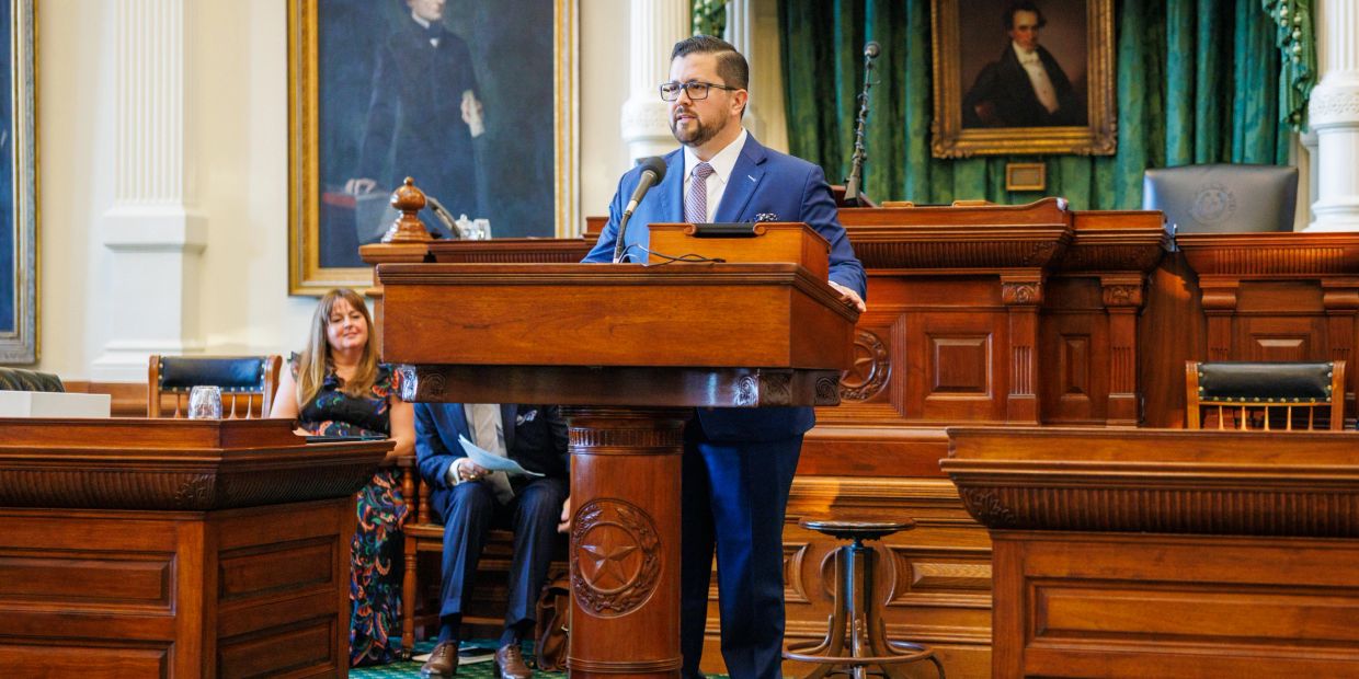 Man in blue suit speaking at a wooden podium in a formal room.