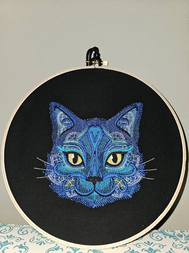 Intricate blue cat embroidery on black fabric in a wooden hoop.