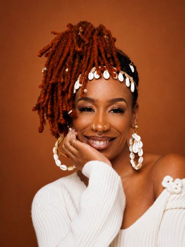 Woman with styled red dreadlocks and shell accessories smiling warmly.