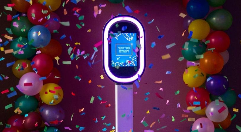 A photo booth surrounded by colorful balloons and confetti with a 'Tap to Start' screen.