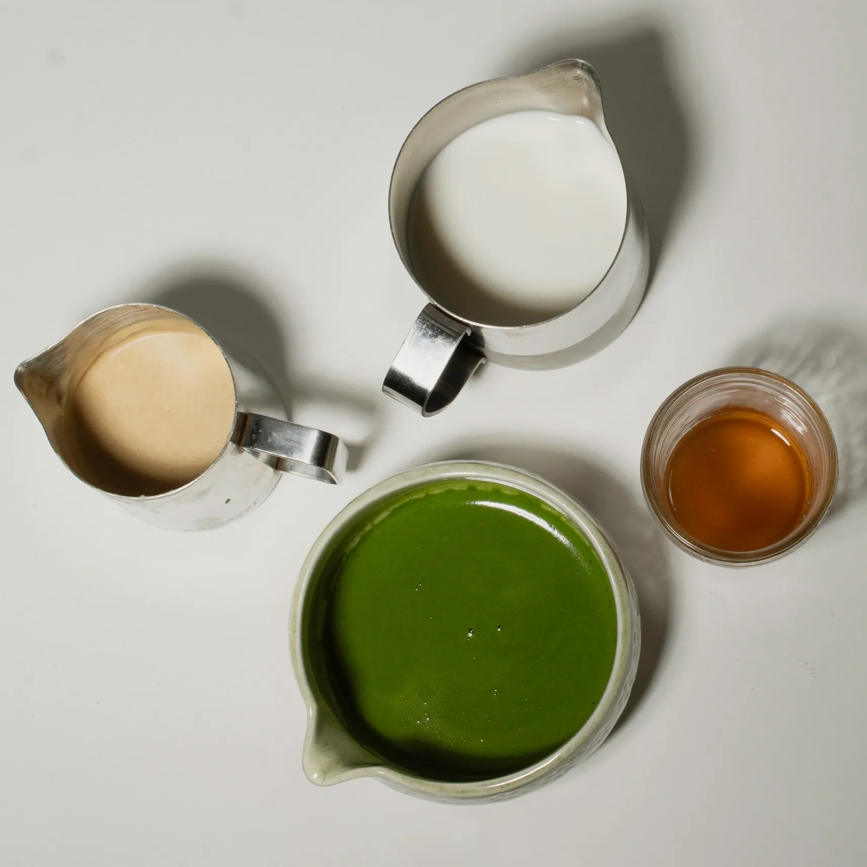 Four containers with green tea, milk, coffee creamer, and honey on a white surface.